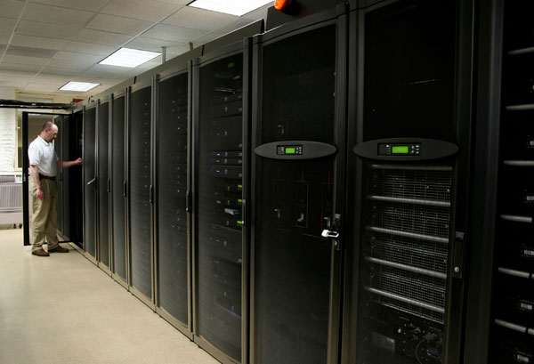 IT technician in a white shirt stands by a row of tall black server racks in a data center corridor, door open to a cabinet.
